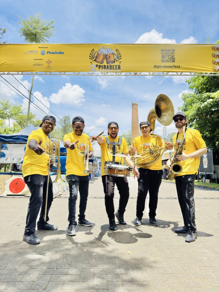 Cinco músicos vestindo camisetas amarelas e com boné escuro, segurando instrumentos musicais, estão posando para fotos em um calçadão.