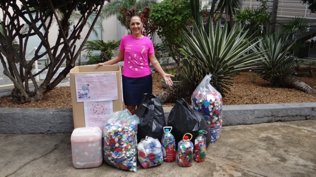 A imagem mostra uma mulher em um ambiente externo, possivelmente um jardim ou área verde, posando ao lado de uma grande quantidade de tampas plásticas recicladas. Ela está vestindo uma camiseta rosa com um laço também rosa, símbolo associado à conscientização sobre o câncer de mama, e uma saia escura. Ao seu redor, há diversas sacolas e recipientes transparentes cheios de tampas plásticas coloridas, além de uma caixa de papelão com cartazes e informações sobre a arrecadação dessas tampas. No cartaz fixado na caixa, lê-se em português: "ARRECADAÇÃO DE TAMPAS PLÁSTICAS", seguido do nome do projeto "Projeto Mãos Amigas" e a frase "O Câncer Une a Amizade". A palavra "FORTALEZA" indica que a ação acontece nessa cidade. A imagem transmite uma mensagem de solidariedade e engajamento comunitário na luta contra o câncer, utilizando a coleta de tampas plásticas como forma de apoio ou arrecadação de fundos. A mulher parece estar orgulhosa e motivada com a iniciativa.