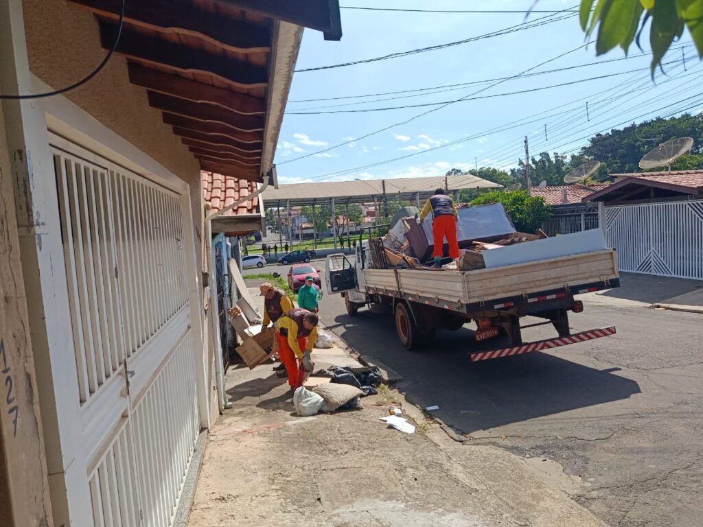 Um grupo de trabalhadores carrega móveis e outros objetos em um caminhão em uma rua de um bairro. A imagem mostra uma rua de um bairro sob um céu claro e ensolarado. No lado esquerdo, parte de uma casa bege com um portão branco é visível. Três homens, vestindo calças laranja e jaquetas com detalhes em amarelo e preto, recolhem objetos e levam para dentro da carroceria de um caminhão branco. Um quarto homem está em cima da carroceria, organizando os objetos que incluem móveis e outros materiais. O caminhão está estacionado na rua, que parece estar numa área residencial. Ao fundo, é possível ver outras casas, árvores e uma estrutura que parece ser um mercado ou feira coberta. Há também carros estacionados ao longo da rua. A atmosfera geral é de trabalho e organização, com os trabalhadores a realizarem a remoção de entulhos ou móveis usados.