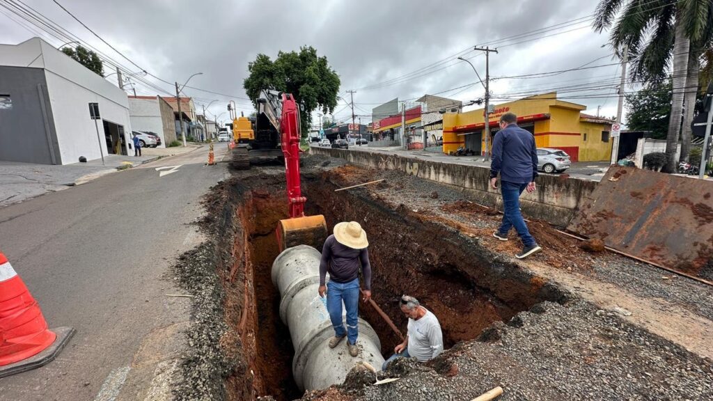A imagem mostra uma cena de construção em uma rua urbana sob um céu nublado. No centro, há uma vala profunda escavada no asfalto, onde dois grandes tubos de concreto estão sendo instalados. Um trator escavador amarelo com a parte da pá em vermelho está posicionado na extremidade da vala, com o braço estendido para dentro dela. Dentro da vala, dois trabalhadores estão presentes: um está em cima de um dos tubos de concreto usando chapéu de palha, camisa roxa e calça jeans, e outro está agachado no fundo da vala, vestindo camiseta branca. Na lateral direita da vala, um homem de jaqueta azul escura e calça jeans caminha próximo à borda. Ao redor, há cones de trânsito, carros estacionados e algumas construções comerciais, incluindo um prédio amarelo com detalhes em vermelho. No canto esquerdo da imagem, há uma placa com os dizeres "LAVA RAPIDO Peixoto Pneus e Rodas". A luz é difusa devido ao céu nublado, e a cena transmite um ambiente de trabalho em uma obra de infraestrutura urbana.