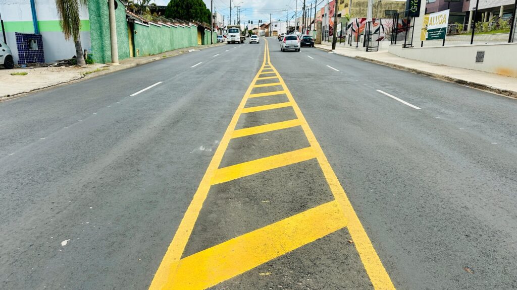 A imagem mostra uma rua urbana larga e pavimentada, com uma faixa amarela no meio que serve como ilha de separação entre as duas pistas de tráfego. A faixa amarela tem marcações em forma de chevron, que indicam para os veículos que não devem ultrapassar aquela área. De um lado da rua, há um muro verde, e do outro lado, há calçadas com postes de iluminação e algumas árvores. Mais ao fundo, é possível ver alguns carros e caminhões transitando pela via. No lado direito da imagem, há uma placa com a palavra "ALUGA", indicando que algum imóvel ou espaço está disponível para locação. O ambiente parece ser um bairro residencial ou comercial em uma cidade, com construções e postes ao longo da rua. O céu está claro, sugerindo que a foto foi tirada durante o dia. A perspectiva da imagem é em ângulo baixo, o que destaca a faixa amarela no centro da rua e dá profundidade à cena.