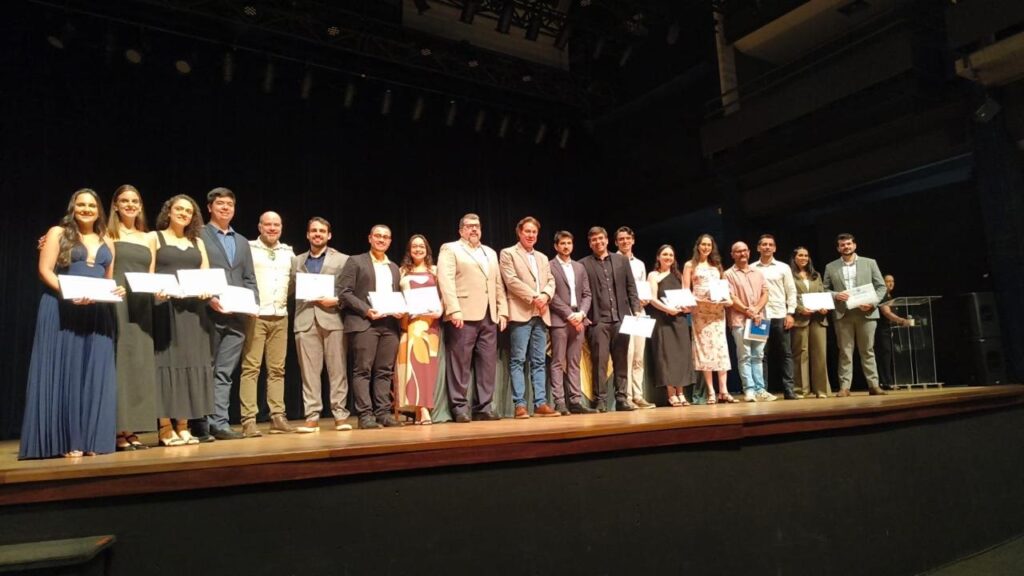 A imagem mostra um grupo de cerca de 20 pessoas alinhadas lado a lado sobre um palco de madeira, possivelmente durante uma cerimônia de formatura ou entrega de certificados. As pessoas, que incluem homens e mulheres de diferentes idades, estão vestidas de forma semi-formal a formal, com ternos, blazers e vestidos. Muitos seguram certificados ou pastas brancas nas mãos. Atrás deles, há uma cortina preta fechada que cobre o fundo do palco. A iluminação está focada no grupo, destacando-os contra o fundo escuro. À direita da imagem, é possível ver um púlpito transparente, sugerindo que o evento pode envolver discursos. O ambiente transmite uma atmosfera de celebração e formalidade.