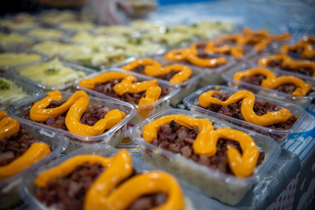 A imagem mostra várias porções individuais de comida dispostas em fileiras organizadas sobre uma mesa. Cada porção está em um recipiente plástico transparente e contém uma base cremosa, possivelmente purê de batata ou arroz, coberta com pedaços pequenos de carne e finalizada com um molho laranja, que parece ser um molho de queijo cheddar. Ao fundo, é possível ver uma pessoa usando luvas plásticas, indicando que a comida está sendo preparada ou servida em um ambiente profissional, como uma cozinha comercial ou serviço de buffet. A mesa está coberta por uma toalha com estampa colorida e há a inscrição "Sweet Food" visível em um dos lados. A imagem transmite a ideia de um serviço de alimentação em grande escala, como catering ou preparo de refeições para entrega.