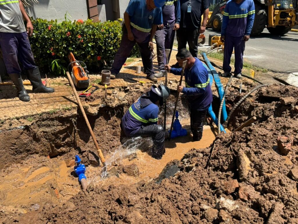 A imagem mostra um grupo de trabalhadores realizando reparos em um cano de água em um grande buraco aberto no chão, que está cheio de lama e água. Dois trabalhadores estão dentro da vala, molhados e cobertos de lama, tentando consertar o cano de onde sai água com pressão. Eles usam uniformes azuis com faixas refletivas. Ao redor da vala, outros trabalhadores observam ou auxiliam. Há ferramentas no chão, como uma serra elétrica e pás, além de mangueiras azuis. Ao fundo, um trator amarelo da marca John Deere está posicionado próximo ao local do reparo. O cenário é ao ar livre, provavelmente em uma rua ou calçada, com vegetação ao fundo. A situação sugere um conserto emergencial de um cano estourado.
