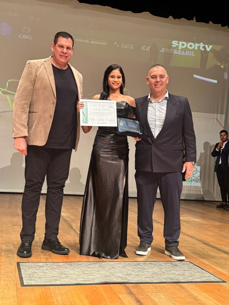 A imagem mostra uma cerimônia de premiação realizada em um palco com piso de madeira. No centro, três pessoas posam para a foto: um homem alto à esquerda, vestido com blazer bege, camiseta preta e calça preta; uma mulher no meio, usando um vestido longo e elegante preto com fenda, segurando um certificado emoldurado; e um homem à direita, usando um terno azul escuro e camisa clara, segurando uma placa preta. Todos estão sorrindo. Ao fundo, há uma tela grande com vários logotipos, incluindo "CBG", "RIO DE JANEIRO", "Loterias CAIXA" e "sportv". Também é possível ver um homem à direita, parcialmente fora do foco, que parece estar aplaudindo. O ambiente é iluminado de forma profissional, típico de eventos formais. O certificado que a mulher segura tem o nome "NICOLE PIRCIO". A cena transmite um momento de reconhecimento e homenagem, possivelmente relacionado ao esporte, conforme indicam as palavras visíveis na tela.