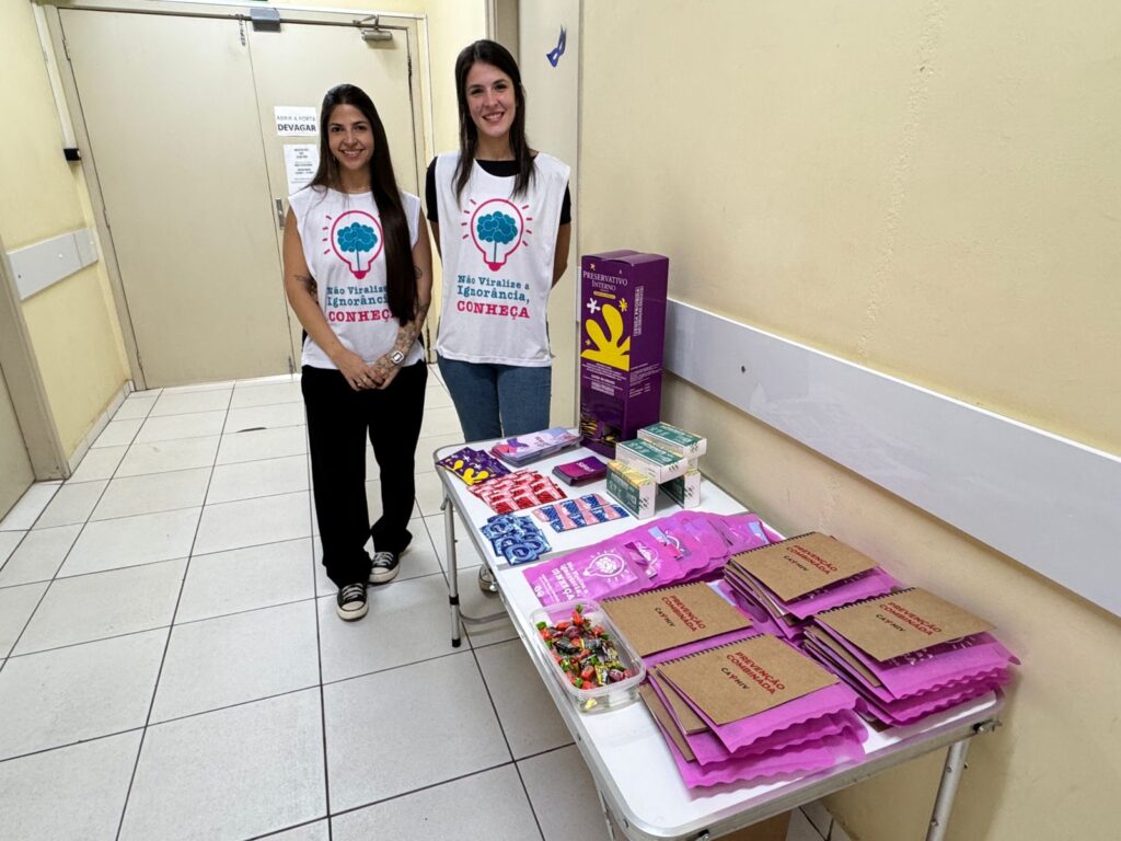 A imagem retrata duas mulheres em um corredor, posicionadas ao lado de uma mesa repleta de materiais informativos, possivelmente promovendo uma campanha de conscientização. A cena é organizada com as duas mulheres em pé lado a lado, ligeiramente descentralizadas para a esquerda, equilibrando a composição com a mesa e os materiais à direita. Elas parecem estar posando para a câmera, criando uma conexão visual direta com o espectador. A ação é estática, capturando um momento de apresentação ou demonstração. A mesa está repleta de panfletos, embalagens e outros materiais promocionais, com uma caixa vertical ao fundo, sugerindo que o evento ou iniciativa está em andamento. O enquadramento é amplo, mostrando o corredor e a porta ao fundo, proporcionando uma perspectiva clara do ambiente. As duas mulheres são os focos principais da imagem. Ambas usam camisetas brancas com um desenho de um cérebro com uma lâmpada e a frase "Não viralize a ignorância, CONHEÇA". A mulher à esquerda tem cabelo castanho escuro e veste calças pretas e tênis. Ela tem uma tatuagem no braço esquerdo e usa um relógio de pulso. A mulher à direita tem cabelo castanho claro e usa calças jeans e uma camiseta preta por baixo da branca. Ambas as mulheres sorriem para a câmera, transmitindo uma sensação de profissionalismo e simpatia.