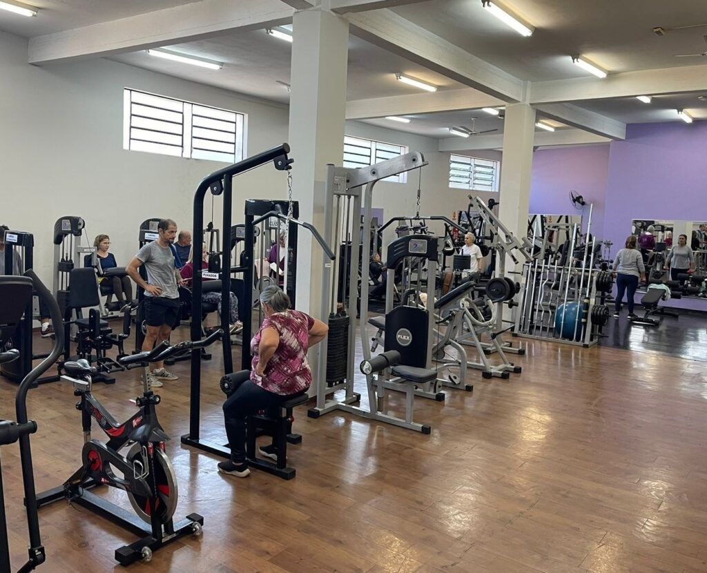 A imagem mostra uma academia interna, bem iluminada e espaçosa, com várias pessoas se exercitando. O ambiente é moderno, com paredes claras e um detalhe em roxo ao fundo. O piso tem aparência de madeira clara, e o teto possui luminárias retangulares que proporcionam uma iluminação uniforme. No primeiro plano, uma mulher está sentada em um aparelho de puxada (lat pulldown), usando uma camiseta rosa com estampa e calça legging preta. Ao lado dela, um homem de camiseta cinza e shorts pretos está em pé, olhando para a frente, com as mãos na cintura. Ao fundo, outras pessoas utilizam diferentes equipamentos, como uma máquina de leg press e uma de cabo. Há também uma parede com espelhos, refletindo o ambiente e os praticantes. Vários aparelhos de musculação estão organizados em filas, incluindo um suporte com barras (halteres) e uma bola azul de exercícios. No canto inferior esquerdo, um equipamento semelhante a uma bicicleta ergométrica é visível. O ambiente transmite uma atmosfera típica de academia, voltada para exercícios de musculação e condicionamento físico.