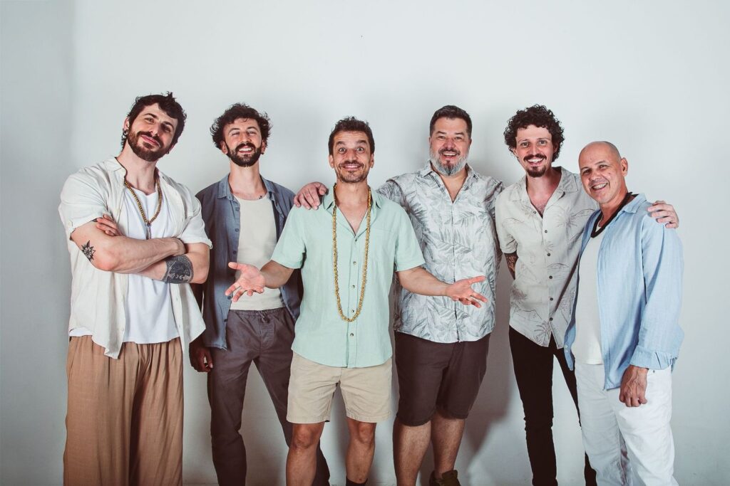 A imagem mostra um grupo de seis homens alinhados lado a lado, posando para a foto contra uma parede branca lisa. Eles estão vestidos de forma casual, com camisas de manga curta ou comprida e calças ou bermudas em tons claros e neutros. Alguns usam acessórios como colares de contas longos. A maioria tem barba e cabelos de diferentes estilos, e um deles é careca. Eles estão sorrindo e parecem estar descontraídos e amigáveis, com alguns com os braços cruzados e outros com os braços ao redor dos ombros dos colegas. A iluminação é clara e uniforme, destacando os rostos e criando um ambiente acolhedor e alegre.