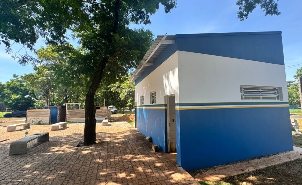 A imagem mostra um pequeno edifício moderno, retangular, com um esquema de cores em azul, branco e amarelo. O telhado é inclinado e feito de metal. Nas paredes do edifício, há várias janelas pequenas com venezianas horizontais, que provavelmente permitem ventilação. Uma porta está visível na lateral do prédio voltada para a frente. O edifício está situado sobre um piso de blocos intertravados, formando uma área pavimentada. À esquerda do edifício, há uma grande árvore com muitas folhas que proporciona sombra sobre parte do piso. Próximo à árvore, há alguns bancos de concreto espalhados pela área pavimentada, sugerindo um espaço para descanso ou espera. Ao fundo, é possível ver outras árvores, um contêiner azul com a inscrição "Locadora de Contêineres" em branco, e um caminhão branco estacionado. A iluminação é forte e indica que a foto foi tirada em um dia ensolarado. Não há pessoas na imagem. No geral, o edifício parece ser uma construção recente ou bem conservada, possivelmente destinada a uso público, como um banheiro, guarita ou espaço de apoio para alguma atividade. A combinação de cores e o entorno bem cuidado dão uma impressão de organização e funcionalidade.