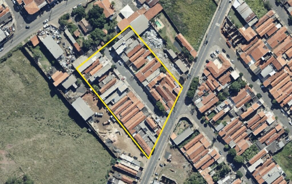 A imagem mostra uma vista aérea de uma área urbana, com foco em um quarteirão retangular destacado por uma borda amarela. Dentro desse quarteirão, há duas fileiras de casas com telhados de cerâmica laranja, muito próximas umas das outras, separadas por uma rua estreita no centro. Ao redor do quarteirão, há ruas pavimentadas com vários carros estacionados, além de algumas construções maiores, possivelmente industriais ou comerciais, com telhados cinza. À esquerda da imagem, há uma área verde aberta, que parece ser um campo ou terreno não urbanizado. A iluminação é forte, indicando que a foto foi tirada durante o dia, com sombras curtas e definidas. A imagem mostra claramente a transição entre uma área residencial densa e áreas mais abertas ou industriais ao redor.