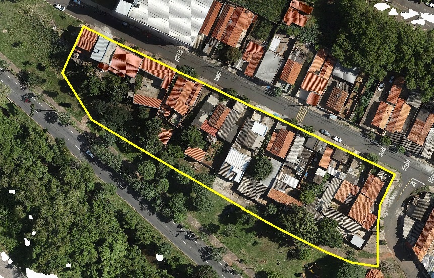 A imagem mostra uma vista aérea de um bairro residencial, com várias casas alinhadas em um terreno estreito e alongado, que está destacado por um contorno amarelo. As casas possuem telhados de cerâmica na cor laranja e alguns com telhados de metal cinza. O terreno está delimitado por ruas asfaltadas, uma delas com uma faixa pintada com a palavra "PARE", indicando uma parada obrigatória no trânsito. À esquerda da área destacada, há um grande espaço verde com muitas árvores, e uma via larga de dois sentidos. À direita, há uma rua mais estreita onde alguns carros estão estacionados. A iluminação é forte, sugerindo que a foto foi tirada durante o dia, com sombras curtas projetadas pelos edifícios e árvores. O ambiente parece ser de uma região urbana em um país de língua portuguesa, provavelmente Brasil.