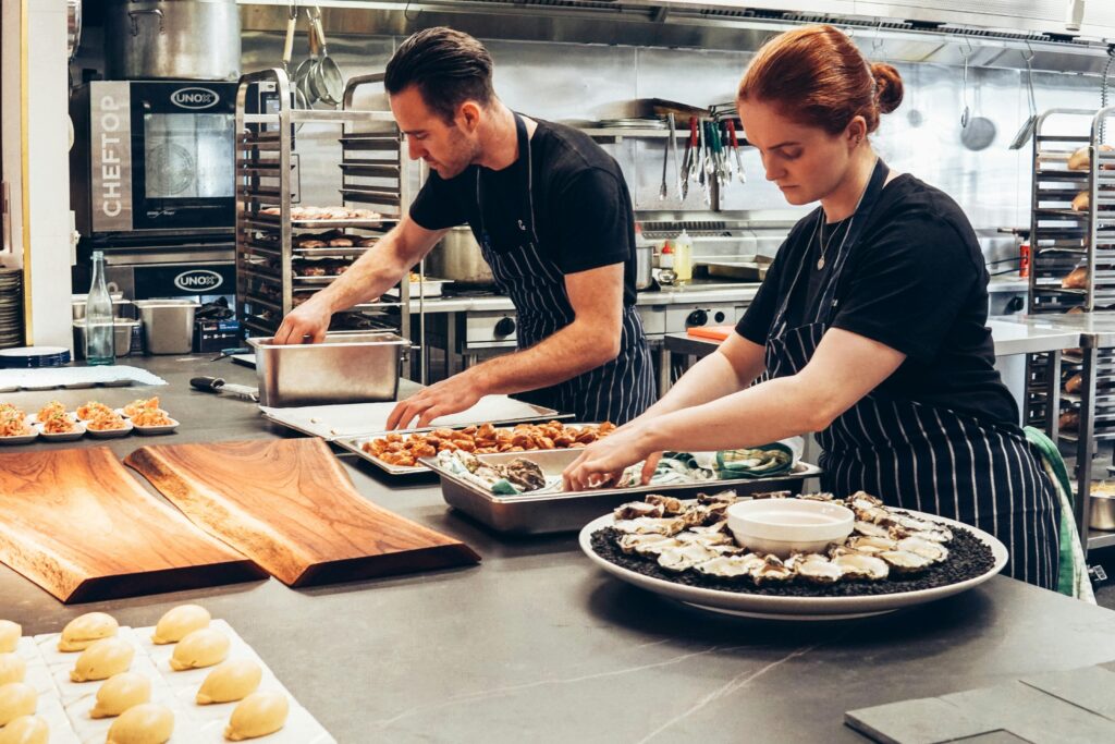 A imagem mostra uma cena em uma cozinha profissional vista de cima. Dois chefs, um homem e uma mulher, estão concentrados preparando e montando pratos sobre uma bancada longa e escura. Ambos usam aventais listrados e camisetas pretas. O homem tem cabelo escuro e barba, enquanto a mulher tem cabelo ruivo preso. Eles estão organizando diferentes tipos de alimentos, incluindo ostras dispostas sobre pedras pretas em um prato grande e circular, além de pequenas porções de aperitivos e uma bandeja com pães ou bolinhos. Ao fundo, é possível ver equipamentos de cozinha industrial, como fornos, prateleiras com utensílios e assadeiras. A iluminação é clara, destacando os detalhes dos alimentos e o ambiente limpo e organizado. A atmosfera transmite profissionalismo e dedicação na preparação culinária.