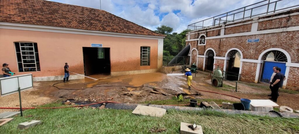 A imagem mostra uma cena de limpeza após uma enchente em um local industrial ou histórico. Dois trabalhadores estão usando mangueiras de alta pressão para remover uma camada espessa de lama marrom que cobre o chão e as paredes inferiores de dois edifícios. À esquerda, há uma construção de cor pêssego com telhado de telhas, e à direita, um prédio de tijolos vermelhos com janelas arqueadas e uma placa escrita "AQUEDUTO 1887". No fundo, é possível ver um grande tubo preto. Algumas pessoas estão observando a limpeza. O chão está completamente coberto por lama e detritos, evidenciando que houve uma inundação recente. O céu está parcialmente nublado, indicando que o tempo já melhorou após o evento. A imagem transmite o esforço para restaurar o local depois do desastre natural.