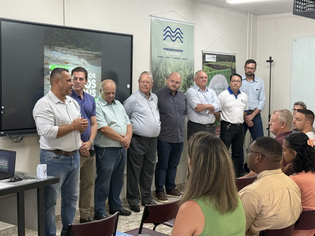 A imagem mostra uma reunião formal em um ambiente interno, possivelmente um escritório ou sala de conferências. O foco principal é um grupo de oito homens alinhados na frente da sala, todos vestidos de forma casual ou semi-formal, com camisas sociais e calças. Um dos homens, à esquerda, está falando, gesticulando com as mãos, enquanto os demais observam atentamente. Ao fundo, há uma grande tela digital exibindo a imagem de uma cachoeira, junto a um banner com o texto "PROGRAMA MUNICIPAL DE PAGAMENTO POR SERVIÇOS AMBIENTAIS", "PRESERVANDO O FUTURO", "CUIDAR DO HOJE PARA GARANTIR O AMANHÃ" e "8ª EDIÇÃO", indicando que o evento está relacionado a um programa ambiental municipal. Na frente, há algumas pessoas sentadas, assistindo à apresentação, e uma mesa com um computador portátil e uma caneca branca. A iluminação é fluorescente, e as paredes são claras, criando um ambiente formal e organizado. A atmosfera sugere uma apresentação ou palestra sobre temas ambientais e sustentabilidade.