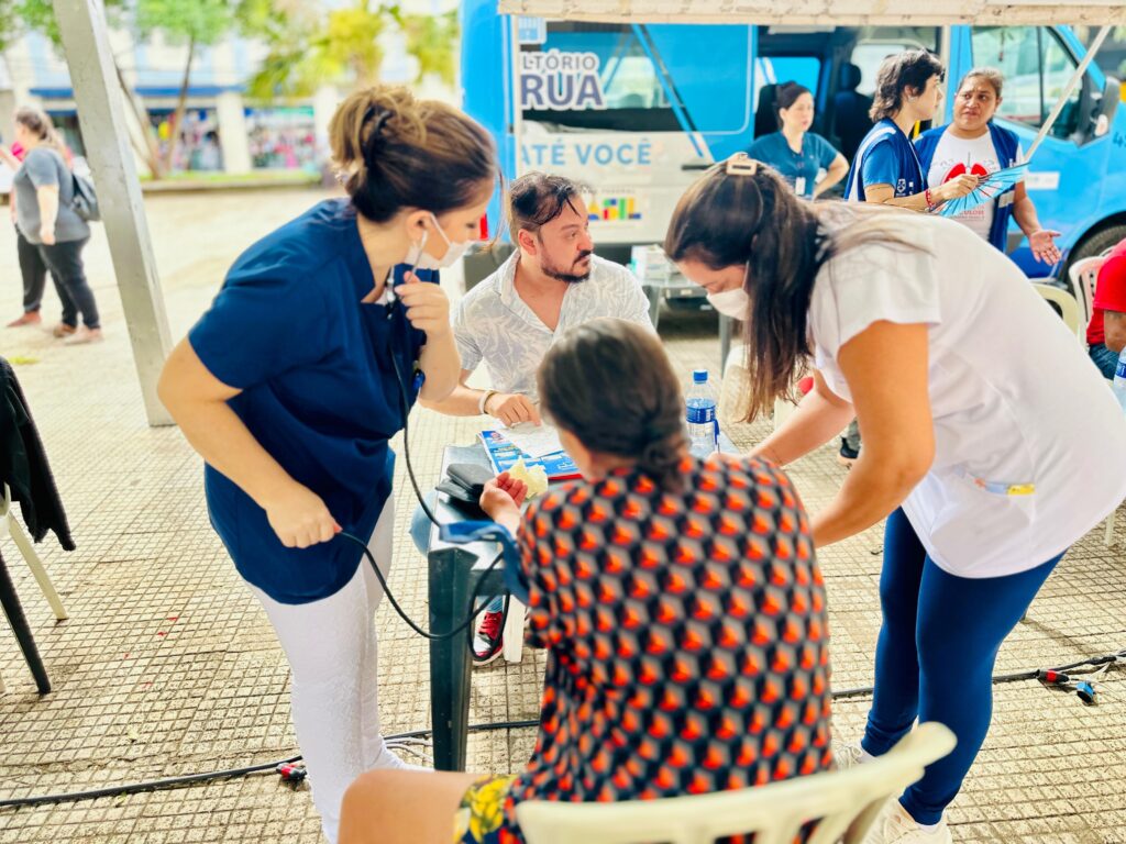 A imagem mostra uma unidade móvel de saúde atuando em um espaço público ao ar livre, possivelmente numa praça de uma cidade. No centro da cena, profissionais de saúde estão atendendo pessoas sentadas em cadeiras ao redor de uma mesa. Uma enfermeira, vestindo um uniforme azul escuro e calça branca, está medindo a pressão arterial de uma paciente que usa uma blusa laranja com estampa preta. Outra profissional, vestindo um uniforme branco e azul, está inclinada sobre a mesa auxiliando no atendimento. Um homem, sentado à mesa, parece estar preenchendo um formulário ou conversando com a equipe. Ao fundo, há uma van azul com a inscrição "CONSULTÓRIO NA RUA ATÉ VOCÊ", indicando que se trata de um serviço móvel de saúde que leva atendimento médico diretamente às pessoas na comunidade. A cena transmite uma ação de cuidado e atenção à saúde em um ambiente comunitário, com a presença de vários profissionais de saúde e pacientes. A iluminação é natural, sugerindo que o atendimento ocorre durante o dia. A composição da imagem destaca o atendimento humanizado e acessível oferecido pelo programa.