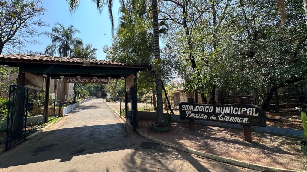 A imagem mostra a entrada do Zoológico Municipal chamado "Paraíso da Criança". A entrada é composta por uma estrutura com um telhado de madeira e telhas, sob a qual há uma placa pendurada com a palavra "ZOOLÓGICO ENTRADA". Ao lado direito da entrada, há uma grande placa de madeira com o nome completo do zoológico: "ZOOLÓGICO MUNICIPAL Paraíso da Criança". O caminho de entrada é pavimentado e cercado por cercas metálicas, com árvores e vegetação ao redor, incluindo palmeiras, que dão um aspecto natural e tropical ao local. A iluminação é forte, sugerindo que a foto foi tirada durante o dia em um dia ensolarado. A cena transmite uma sensação acolhedora e convidativa para os visitantes do zoológico.