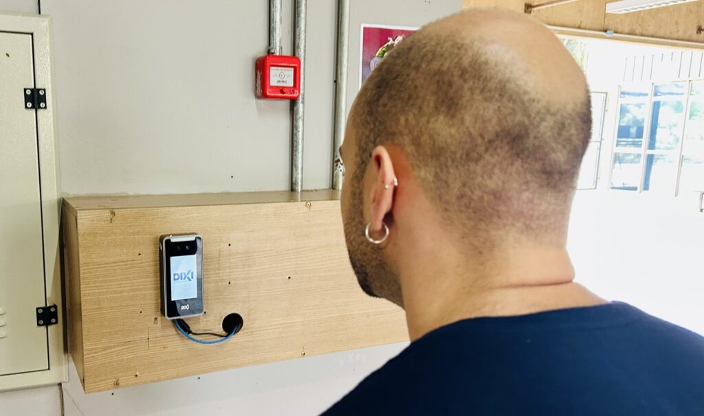 A imagem mostra um homem visto de costas, com a cabeça raspada, olhando para um dispositivo eletrônico fixado em uma parede de madeira clara. O dispositivo parece ser um aparelho de reconhecimento facial ou um relógio de ponto digital, com a marca "DIXI" exibida na tela. Acima do dispositivo, há uma caixa vermelha de alarme de incêndio com a inscrição "ALARME DE INCÊNDIO". O ambiente é interno, com iluminação clara, e ao fundo é possível ver uma porta aberta que leva a outro espaço iluminado por luz natural. O homem está vestido com uma camiseta azul escura e usa dois brincos na orelha esquerda. A cena sugere um local de trabalho ou uma área de controle de acesso, onde o homem utiliza o aparelho para registrar sua entrada ou saída.