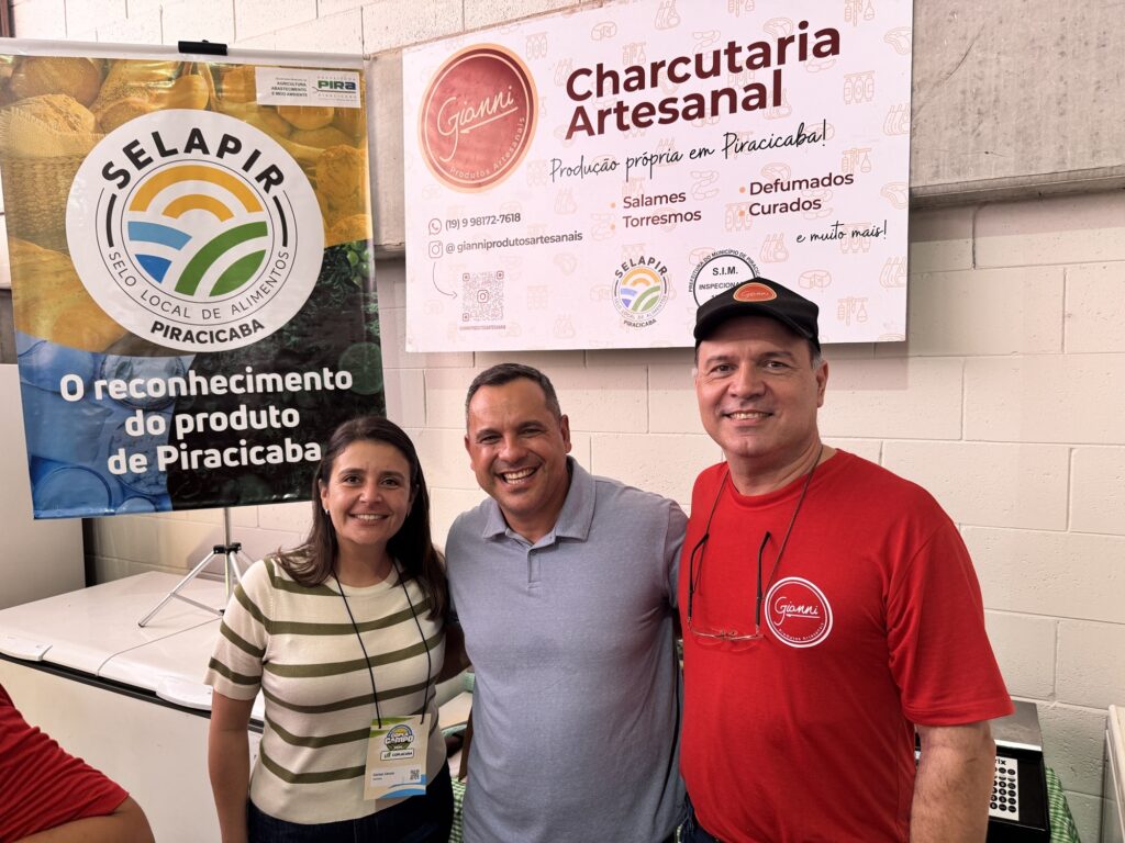 A imagem mostra três pessoas posando para uma foto em um ambiente interno, provavelmente em um evento ou feira de alimentos artesanais. À esquerda, uma mulher com cabelo longo e escuro, usando uma camiseta listrada verde e branca e um crachá no pescoço, sorri para a câmera. No centro, um homem de camiseta polo azul claro também sorri de forma amigável. À direita, outro homem usa camiseta vermelha com o logo da marca "Gianni" e um boné preto, além de usar óculos pendurados na camisa. Ao fundo, há dois banners promocionais. O banner à esquerda exibe o selo "SELAPIR", que representa o reconhecimento de produtos locais de Piracicaba. O banner à direita é da marca "Gianni Produtos Artesanais", que destaca a charcutaria artesanal produzida em Piracicaba, listando produtos como salames, torresmos, defumados e curados, além de informações de contato e redes sociais. O cenário tem uma parede branca e uma geladeira branca ao fundo, sugerindo um ambiente organizado e profissional. A atmosfera geral é positiva, com foco na divulgação de produtos locais e artesanais.