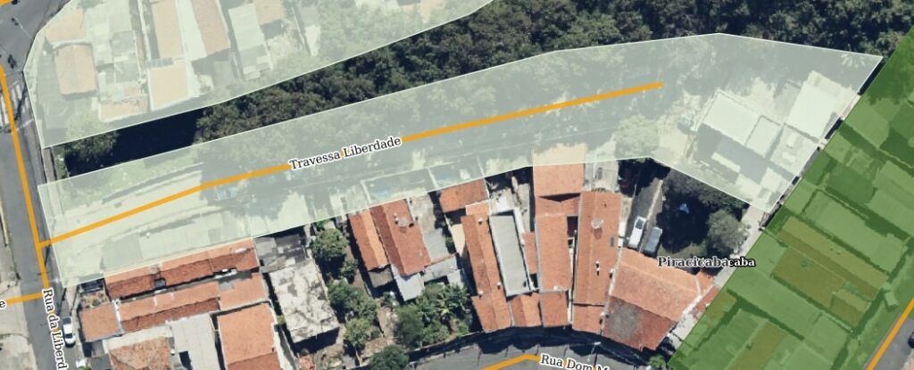 A imagem mostra uma vista aérea de um bairro residencial com ruas, casas e uma área de vegetação densa. No centro da imagem, há uma rua chamada "Travessa Liberdade" destacada com uma sobreposição branca translúcida. Abaixo dessa rua, há várias casas com telhados de cor laranja-avermelhada, muito próximas umas das outras, formando um conjunto residencial compacto. À esquerda, há outra rua chamada "Rua da Liberd" (aparentemente abreviação de Liberdade) que corre verticalmente. Na parte superior direita, uma grande área verde com muitas árvores indica uma zona de vegetação ou mata. No canto inferior direito, aparece a inscrição "Piracicabacaba", que provavelmente indica o nome do bairro ou região. Também é possível ver parte de outra rua chamada "Rua Dom M". A imagem tem boa iluminação, típica de fotos de satélite em dias ensolarados, e mostra sombras nítidas das construções e árvores.