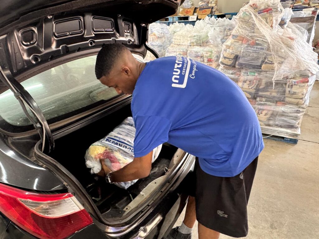 A imagem mostra um jovem homem negro vestindo uma camiseta azul com a inscrição "nutre CESTAS BÁSICAS Compra". Ele está carregando um grande saco plástico transparente, contendo alimentos, para dentro do porta-malas de um carro escuro. O saco parece conter diversos itens básicos de alimentação, como parte de uma cesta básica. Ao fundo, é possível ver várias paletes empilhadas com mais sacos similares, todos envoltos em plástico transparente, indicando um ambiente de depósito ou centro de distribuição. O cenário é interno, com piso de concreto, sugerindo uma área de armazenamento ou logística. O homem está concentrado na tarefa, usando também um relógio e shorts pretos com o logo da Lacoste. A imagem transmite uma ação relacionada à distribuição ou compra de cestas básicas, provavelmente em um contexto de auxílio alimentar.