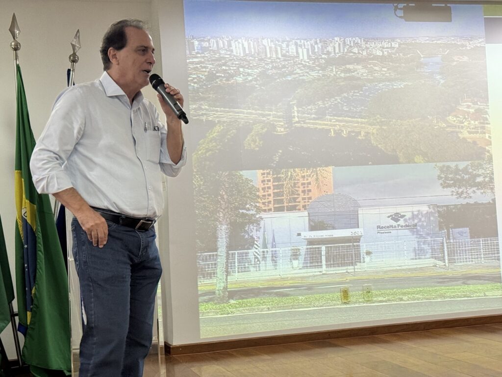 A imagem mostra um homem de meia-idade, com cabelo escuro, vestido com uma camisa azul clara e calça jeans azul escura, falando em um microfone de mão preto. Ele está em pé, com a mão direita no bolso, olhando para a direita, aparentemente fazendo uma apresentação. Ao seu lado, há dois mastros com bandeiras, sendo uma delas a bandeira do Brasil. Ao fundo, há uma grande tela de projeção exibindo duas imagens: uma vista aérea de uma cidade com um rio e pontes, e outra de um edifício moderno com a placa "Receita Federal". O ambiente parece ser uma sala de conferências ou reunião, com piso de madeira clara e iluminação interna. A cena sugere um evento formal ou uma apresentação relacionada a assuntos governamentais, possivelmente em Piracicaba, Brasil.