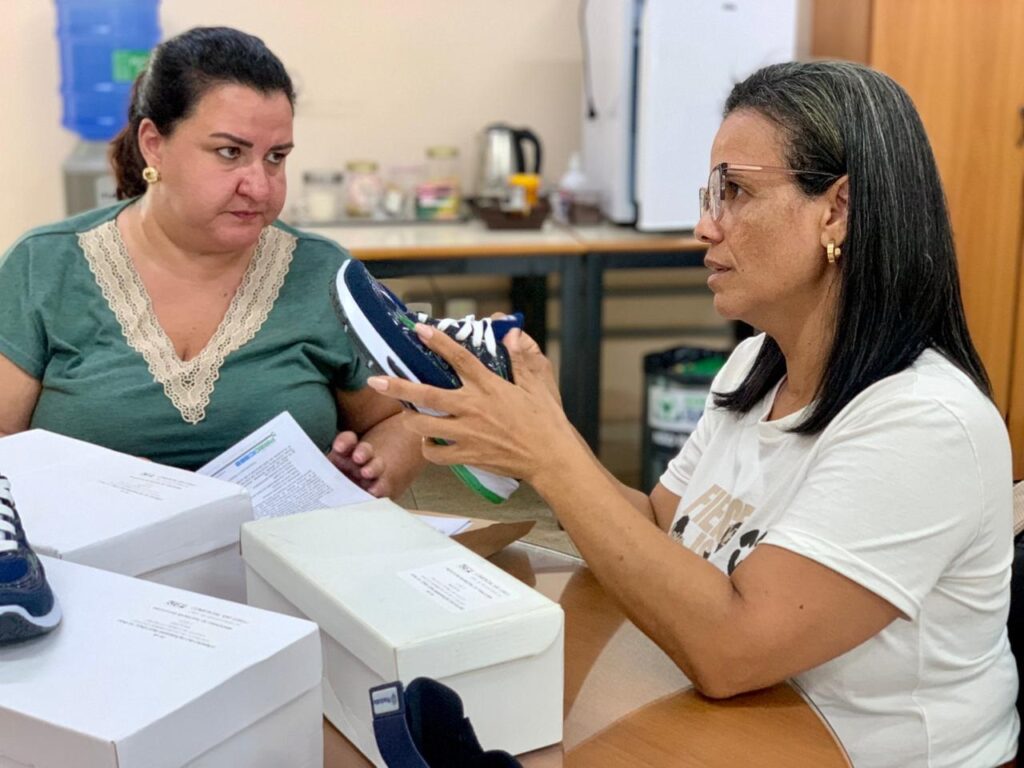 A imagem mostra duas mulheres sentadas à mesa em um ambiente que parece ser um escritório ou local de atendimento. A mulher à direita, que usa óculos e uma camiseta branca, está segurando e examinando um tênis esportivo azul marinho com cadarços brancos e sola verde. A mulher à esquerda, vestindo uma blusa verde com detalhes em renda, observa o tênis com uma expressão séria ou atenta. Sobre a mesa há várias caixas de tênis brancas, algumas com etiquetas, além de documentos espalhados. Ao fundo, é possível ver um bebedouro com garrafão azul, uma prateleira com potes e utensílios, e uma geladeira branca. A iluminação é clara, destacando a interação entre as duas mulheres e o produto que estão analisando. A cena transmite a ideia de uma avaliação ou conferência de materiais, possivelmente em um contexto de doação ou distribuição de calçados.