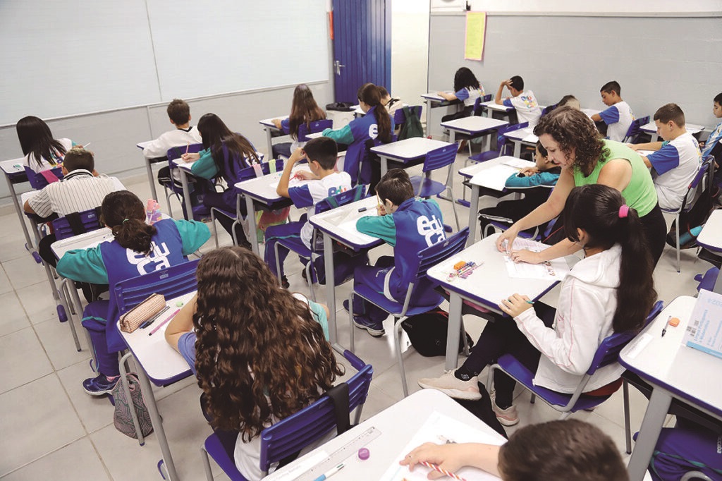 A imagem mostra uma sala de aula com vários alunos sentados em carteiras individuais, todos concentrados em suas atividades, provavelmente escrevendo ou desenhando. A maioria dos estudantes veste uniformes nas cores azul e verde, com a palavra "Educa" estampada nas costas das camisetas. A professora, uma mulher com cabelo cacheado e usando uma blusa verde, está inclinada ajudando um aluno na parte lateral direita da sala. A sala é clara e organizada, com paredes brancas, um quadro branco ao fundo e uma porta azul. O ambiente transmite uma atmosfera de concentração e aprendizado, típica de um ambiente escolar.