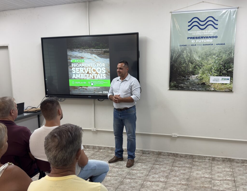 A imagem mostra um homem em pé, aparentemente fazendo uma apresentação ou palestra para um pequeno grupo de pessoas sentadas de frente para ele. Ele veste uma camisa de mangas compridas listrada clara, calça jeans azul e sapatos marrons. O homem está com as mãos entrelaçadas à frente do corpo e sorri levemente, olhando para a audiência. Ao fundo, há uma televisão grande exibindo um cartaz que anuncia a "8ª EDIÇÃO PAGAMENTO POR SERVIÇOS AMBIENTAIS", com uma imagem de uma cachoeira. Ao lado da televisão, há uma faixa vertical pendurada na parede com o texto "PRESERVANDO O FUTURO" e o logo da Prefeitura de Piracicaba. O ambiente parece ser uma sala clara, com paredes brancas, piso de azulejos claros com padrão marmorizado e algumas instalações elétricas aparentes na parede. Na parte inferior da imagem, é possível ver algumas pessoas sentadas, assistindo à apresentação. O cenário transmite um clima profissional e informal, possivelmente relacionado a um evento municipal sobre meio ambiente e agricultura.