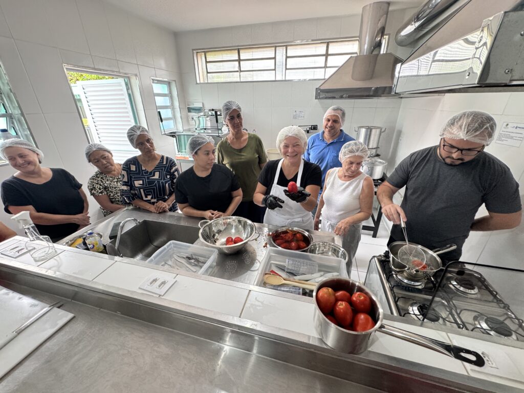 A imagem mostra um grupo de nove pessoas reunidas em uma cozinha industrial bem iluminada, participando de uma aula ou oficina de culinária focada em tomates. No centro da cena, uma mulher com avental branco e luvas pretas segura um tomate descascado, enquanto um homem ao lado dela, também usando luvas, utiliza uma escumadeira para retirar tomates de uma panela fervente em um fogão a gás. As outras pessoas ao redor observam atentamente a demonstração. Todos usam toucas brancas para manter a higiene. A cozinha possui paredes de azulejo branco, bancadas e pias de aço inox, além de um exaustor grande sobre o fogão. Há várias tigelas e panelas com tomates sobre a bancada e o fogão. A atmosfera é de aprendizado e colaboração, e a iluminação é clara, vinda de lâmpadas fluorescentes e possivelmente de uma janela ao fundo. A cena sugere um ambiente de escola de culinária ou centro comunitário durante uma atividade prática de cozinha.