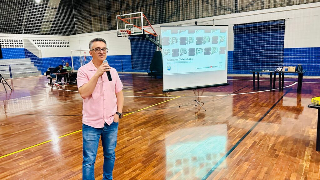 A imagem mostra um homem falando em um microfone dentro de um ginásio esportivo, possivelmente durante uma apresentação ou evento. Ele está vestido com uma camisa rosa clara de mangas curtas e jeans azuis, e usa óculos. Atrás dele, há um telão com um projetor exibindo o título "Programa Cidade Legal", associado à Secretaria de Desenvolvimento Urbano e Habitação da cidade de São Paulo. O ambiente é um ginásio com piso de madeira e marcações para esportes, uma cesta de basquete ao fundo e paredes azuis. À esquerda, duas pessoas estão sentadas em uma mesa com equipamentos, possivelmente controlando o som ou a projeção. A cena sugere uma reunião, palestra ou apresentação pública relacionada a um programa urbano da prefeitura de São Paulo.