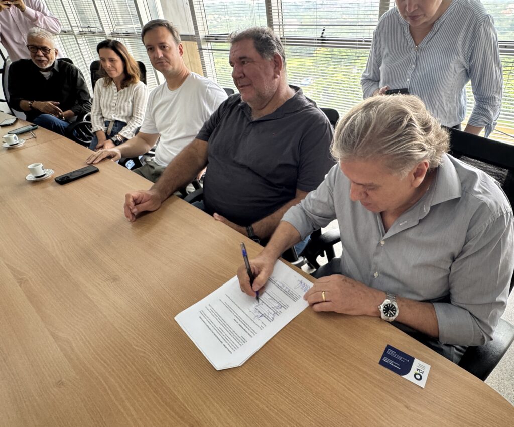 A imagem mostra um ambiente de reunião profissional, possivelmente em um escritório moderno com vista para uma área verde através de grandes janelas. Ao redor de uma mesa de madeira longa, várias pessoas estão reunidas, algumas sentadas e outras em pé. O foco principal é um homem de cabelos grisalhos, vestido com uma camisa azul clara, que está assinando um documento com uma caneta azul. Ao seu lado, outro homem observa atentamente a assinatura. Na mesa, há dois pequenos copos de café, um celular preto e um cartão de visita com o nome "Alexandre Ramos L. Martins" e a sigla "IQA - Instituto de Qualidade Automotiva". O ambiente é iluminado por luz natural, transmitindo uma atmosfera de formalidade e profissionalismo, sugerindo que um acordo ou contrato importante está sendo formalizado.