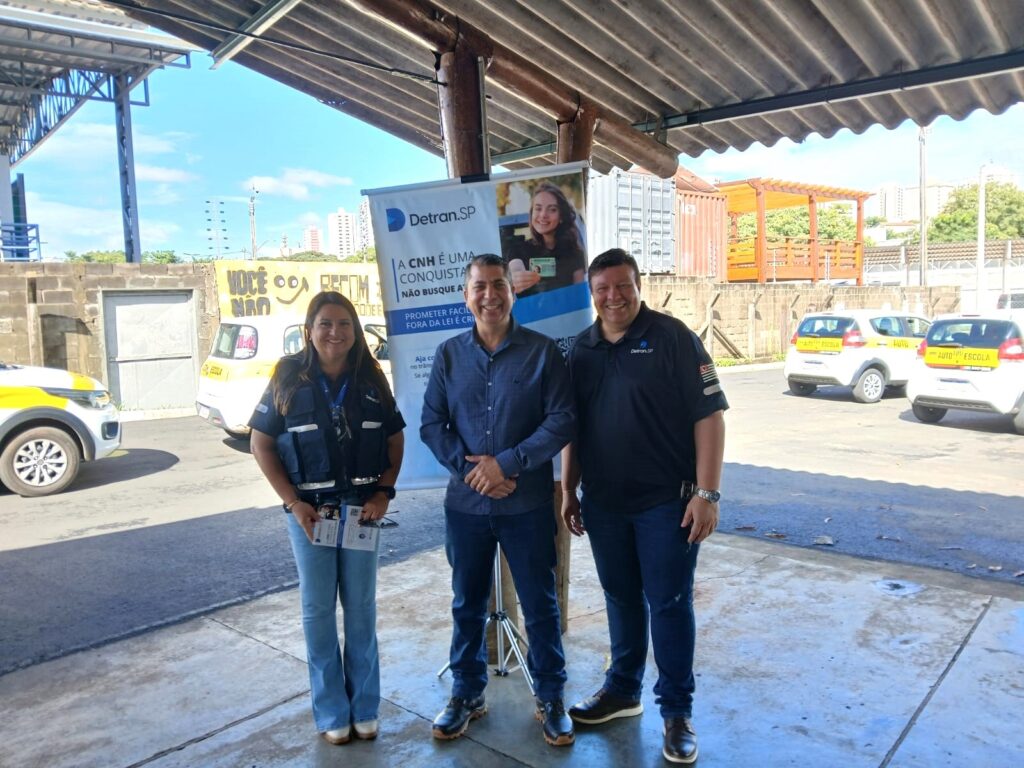 A imagem mostra três pessoas posando para uma foto sob uma estrutura com teto metálico ondulado, provavelmente em um evento ou local do Detran.SP (Departamento de Trânsito de São Paulo). À esquerda, uma mulher com cabelo longo e escuro veste uma camiseta azul clara e um colete preto, segurando folhetos informativos. No centro, um homem de camisa azul escura e jeans sorri para a câmera. À direita, outro homem usa uma camisa polo preta com o logo do Detran.SP e também sorri. Ao fundo, há uma faixa vertical com o logo do Detran.SP e mensagens de conscientização sobre a importância de obter a CNH (Carteira Nacional de Habilitação) de forma legal, alertando contra promessas de facilidade e atalhos ilegais, que são crimes. A faixa traz a frase "A CNH é uma conquista. Não busque atalhos. Prometer facilidade fora da lei é crime. Aja com ética no trânsito. Se alguém te oferecer... VOCÊ NÃO".