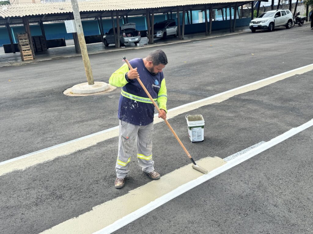 A imagem mostra um homem pintando uma faixa no chão de um estacionamento ou via pavimentada. Ele está usando um rolo grande para aplicar uma tinta de cor clara entre duas linhas brancas já existentes. O homem veste uma camisa azul escura com mangas amarelas fluorescentes e faixas refletivas, calças de trabalho cinza com faixas refletivas e sapatos de segurança marrons. Sua roupa está salpicada de tinta. Ao lado dele, há um balde branco de tinta. Ao fundo, há uma estrutura coberta com teto de metal onde estão estacionados vários carros, incluindo um hatch prata com o porta-malas aberto e um SUV branco. A iluminação é uniforme, indicando um dia nublado. O chão é de asfalto com textura visível. No uniforme do homem, há o logotipo com a palavra "Guaicurus".