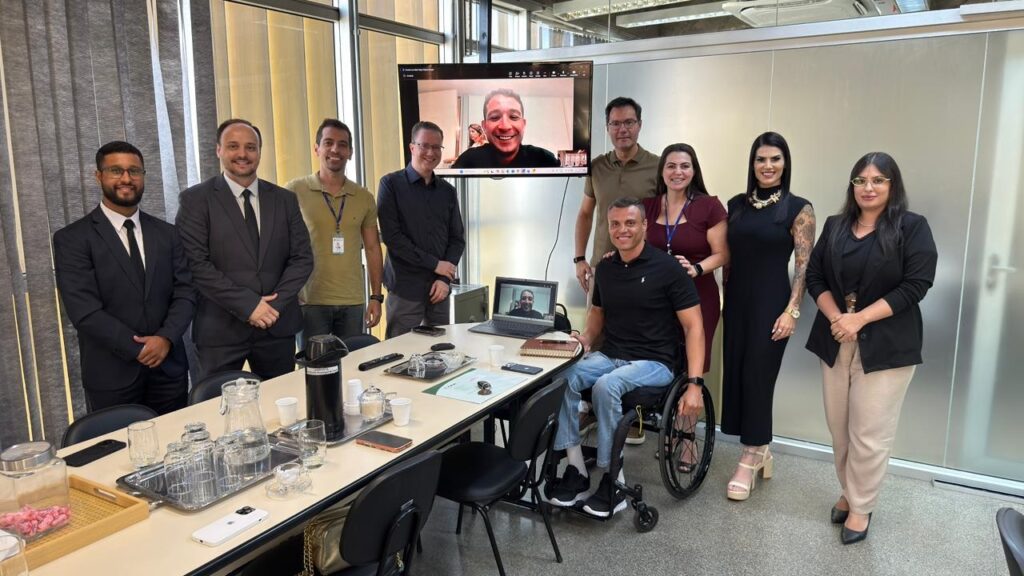 A imagem mostra um grupo de nove pessoas em um ambiente de escritório moderno, provavelmente uma sala de reuniões. Quatro homens estão à esquerda, vestidos formalmente ou casualmente, incluindo um homem em cadeira de rodas no centro, que está sorrindo. À direita, há mais quatro pessoas, incluindo três mulheres e um homem, todos vestidos de forma profissional. Ao fundo, uma grande tela exibe a imagem de um homem participando de uma videoconferência, que também aparece em um laptop sobre a mesa da reunião. A sala tem uma mesa longa com jarros de água, copos e documentos, e o ambiente é bem iluminado, com janelas cobertas por persianas e um teto com acabamento em concreto aparente. A atmosfera geral é profissional e colaborativa.