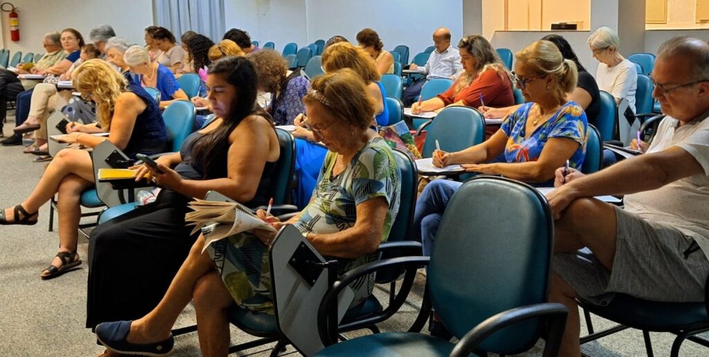 A imagem mostra um grupo de adultos participando de uma aula, palestra ou workshop em uma sala ampla e bem iluminada. As pessoas estão sentadas em cadeiras verdes com pequenas mesas acopladas, organizadas em filas. A maioria está concentrada, escrevendo em cadernos ou lendo, demonstrando atenção ao conteúdo apresentado. Entre os participantes, há uma mulher usando um vestido preto que está com um celular na mão, enquanto outras pessoas ao redor estão focadas em anotações. O ambiente parece ser uma sala de aula ou auditório, com paredes claras e um piso cinza. Ao fundo, é possível ver um extintor de incêndio preso à parede. A atmosfera geral é de estudo e aprendizado, com um público predominantemente adulto e de idade mais avançada.