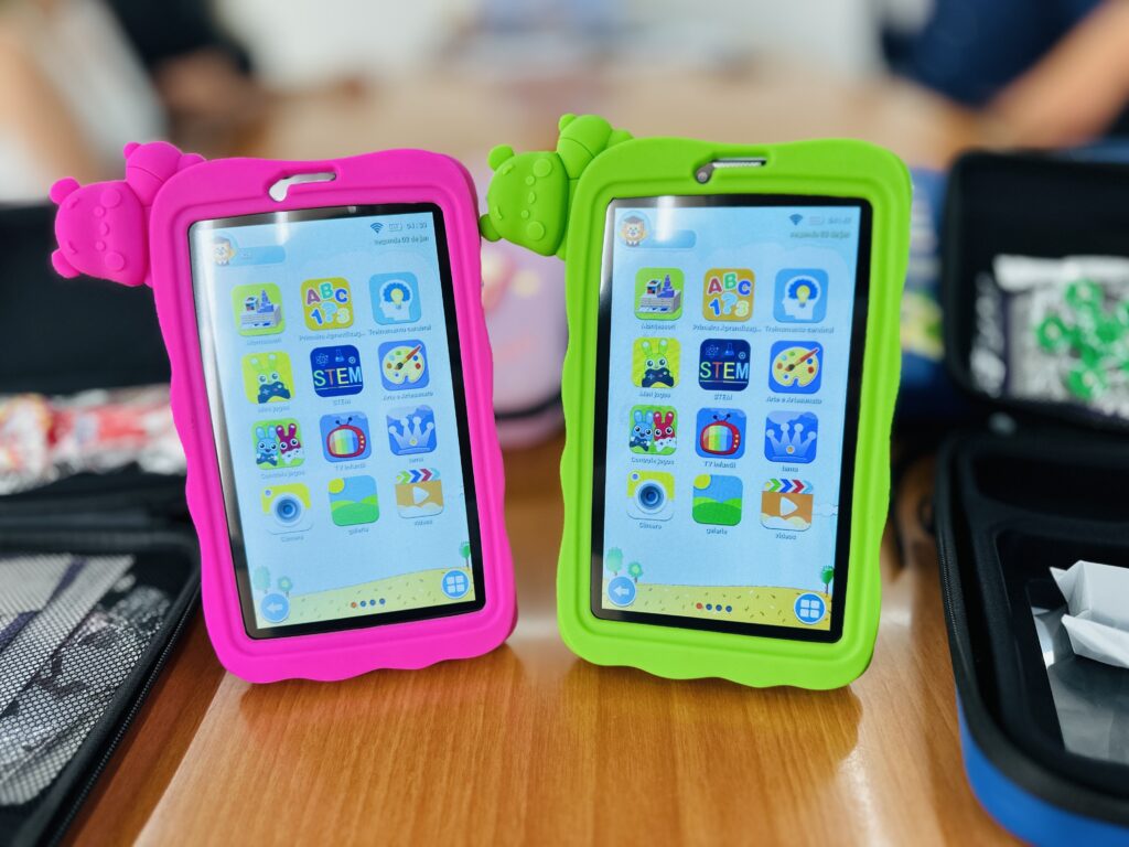 A imagem mostra dois tablets infantis idênticos posicionados lado a lado sobre uma superfície de madeira clara. O tablet à esquerda está protegido por uma capa de silicone rosa vibrante, enquanto o da direita tem uma capa verde-limão igualmente vibrante. Ambas as capas possuem uma figura de personagem de desenho animado, parecendo um ursinho, fixada no canto superior esquerdo. As telas dos tablets estão ligadas e exibem uma interface colorida com ícones grandes e fáceis de identificar, voltados para o público infantil. Os ícones representam diferentes aplicativos e categorias, como "Montessori", "ABC 123", "STEM", "Mini jogos", "Arte e Artesanato", "TV Infantil", "Câmera", "Galeria" e "Vídeos". O fundo da tela mostra um céu azul com algumas nuvens e um campo verde, criando um ambiente amigável e atraente para crianças. No canto superior esquerdo das telas, aparece a hora (04:39 e 04:40) e a data (segunda, 02 de janeiro). O texto está em português, sugerindo que o dispositivo está configurado para esse idioma. Ao fundo, desfocado, é possível ver partes de bolsas ou mochilas pretas e o braço de uma pessoa, indicando que a foto foi tirada em um ambiente interno, possivelmente uma casa ou loja. A imagem tem boa iluminação, destacando as cores vivas das capas e a nitidez da tela dos tablets. O foco está nos dispositivos, com o fundo propositalmente desfocado para dar ênfase aos tablets.