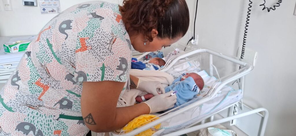 A imagem mostra uma enfermeira cuidando de um recém-nascido em uma unidade hospitalar, provavelmente uma maternidade ou berçário neonatal. A enfermeira, que usa luvas brancas e uma blusa de uniforme com estampas de animais, está inclinada sobre o berço de acrílico onde o bebê está deitado, vestido com uma roupa azul. Ela parece estar realizando um procedimento delicado, como uma coleta de sangue ou exame no calcanhar do bebê. O ambiente é limpo e bem iluminado, com paredes brancas e equipamentos médicos ao redor. Ao fundo, é possível ver outro bebê parcialmente visível em um berço semelhante. A atmosfera transmite cuidado e atenção especializada ao recém-nascido.