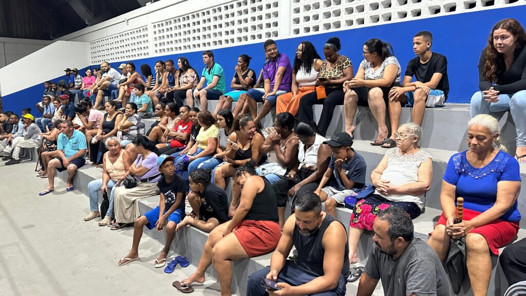 A imagem mostra uma plateia diversificada sentada em arquibancadas de concreto dentro de um ginásio ou centro comunitário. Cerca de 40 a 50 pessoas de diferentes idades e etnias estão reunidas, vestindo roupas casuais de verão, como camisetas, regatas, shorts e chinelos. Algumas pessoas estão atentas ao que acontece fora do quadro, enquanto outras conversam, olham para o celular ou parecem distraídas. O ambiente é iluminado por luzes internas, com uma parede azul ao fundo e uma estrutura branca com aberturas retangulares na parte superior. A foto foi tirada em ângulo amplo, mostrando a disposição das pessoas em diagonal, com as arquibancadas se estendendo da frente para o fundo da imagem. No canto inferior direito, destaca-se uma mulher com blusa azul e saia vermelha segurando um guarda-chuva. A cena transmite uma atmosfera de reunião comunitária ou evento local.