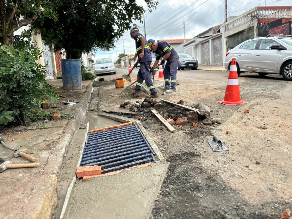 A imagem mostra uma cena de construção em uma rua residencial em um dia nublado. Dois trabalhadores estão no centro da imagem, vestindo uniformes azuis com faixas refletivas amarelas e capacetes de segurança. Eles estão misturando ou mexendo concreto fresco sobre o asfalto. Em primeiro plano, há uma grade de dreno recém-instalada, feita de metal azul, embutida no concreto ainda úmido. Ao redor, há ferramentas espalhadas, como uma colher de pedreiro, um martelo e um tambor azul. À direita, um cone de trânsito laranja e branco está posicionado para sinalizar a obra. Ao fundo, é possível ver carros estacionados e casas de paredes claras, compondo uma típica rua residencial. A imagem é capturada de um ângulo baixo, destacando o trabalho no dreno e criando profundidade na cena.