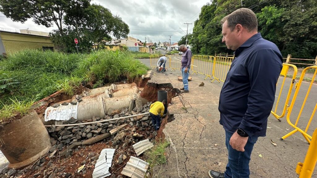 A imagem mostra uma cena de construção ao ar livre em uma rua onde uma parte do asfalto e da calçada desabou, revelando grandes tubos de concreto usados para drenagem subterrânea. No primeiro plano, há um homem vestido com camiseta azul escura e calça jeans olhando para o buraco. No centro, um trabalhador com camiseta amarela e calça azul está agachado dentro da área escavada, aparentemente trabalhando em um tubo preto corrugado. Ao fundo, outros dois trabalhadores aparecem, um de camiseta roxa e jeans próximo a uma barreira metálica amarela, e outro agachado próximo a equipamentos. O chão está coberto por asfalto rachado, terra vermelha, pedras cinzas e detritos. Barreiras metálicas amarelas cercam a área interditada para segurança. Ao fundo, vê-se uma rua com casas residenciais, árvores verdes e um céu nublado. Há uma placa de pare visível à esquerda da imagem. A iluminação é difusa, indicando um dia nublado. A foto foi tirada de um ângulo levemente elevado, destacando a extensão do desabamento e o trabalho dos operários.