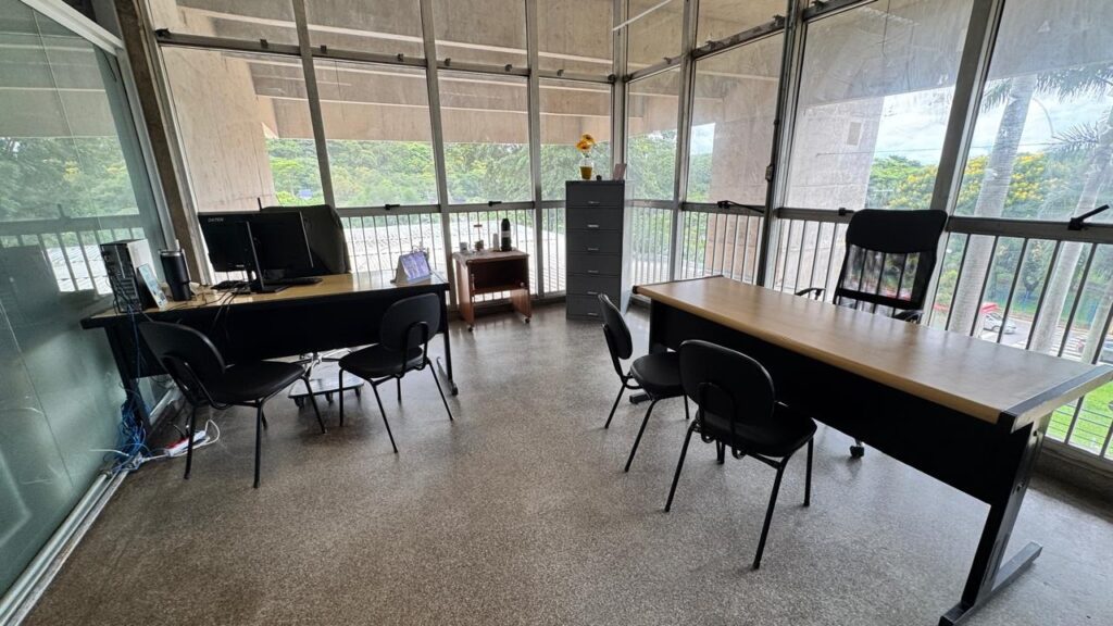 A imagem mostra um escritório moderno e espaçoso, com grandes janelas do chão ao teto que permitem a entrada abundante de luz natural. O ambiente é decorado com uma paleta de cores neutras, predominando tons de cinza, preto e madeira clara. Há duas mesas principais: uma delas possui um computador com monitor, teclado e torre, enquanto a outra está vazia. Várias cadeiras de escritório pretas estão posicionadas próximas às mesas. No canto da sala, há um armário alto cinza e, sobre ele, um vaso com flores amarelas que traz um toque de cor ao ambiente. Também há uma pequena mesa lateral de madeira com um objeto que parece ser um garrafa térmica. O escritório tem um aspecto organizado e profissional, com vista para árvores e palmeiras do lado de fora, sugerindo que o local está em uma região de clima tropical ou subtropical. No monitor do computador, aparece a palavra "DATEN". O espaço parece ideal para atividades administrativas ou reuniões de trabalho.