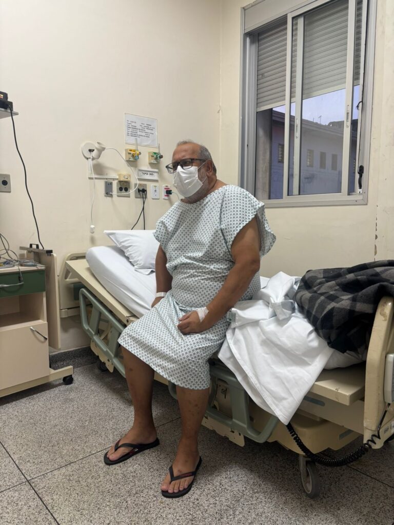 A imagem mostra um homem de meia-idade sentado na beirada de uma cama de hospital. Ele está vestido com uma camisa hospitalar branca com pequenos desenhos azuis, usando máscara facial branca, óculos de aro preto e chinelos pretos. O homem tem um curativo com um cateter intravenoso na mão esquerda e uma pulseira de identificação no pulso direito. O ambiente é um quarto de hospital com paredes bege e piso de azulejo claro. Atrás da cama, há uma janela com grades metálicas e persianas, além de equipamentos médicos fixados na parede, como conexões para oxigênio e sucção, e um papel com informações presas na parede. A cama está arrumada com lençóis brancos e um cobertor xadrez escuro dobrado na ponta. A iluminação é clara e típica de um ambiente hospitalar. O homem parece estar olhando levemente para o lado, com expressão neutra. No canto da imagem, há um pequeno criado-mudo. No texto extraído da imagem, lê-se o nome "Fabio Henrique de Souza", data de nascimento "04/02/2000" e informações possivelmente relacionadas à internação hospitalar, como número de registro, unidade e leito.