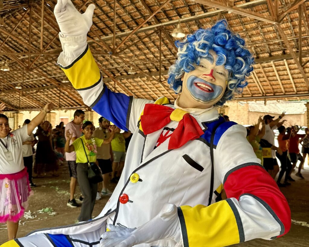 A imagem mostra um palhaço vestido com um traje colorido e vibrante, predominando as cores azul, vermelho, branco e amarelo. Ele usa uma peruca azul cacheada, luvas brancas e uma roupa branca com grandes botões amarelos e vermelhos. O palhaço está sorrindo, com o rosto pintado de branco, nariz vermelho e detalhes azuis ao redor da boca. Ele está em primeiro plano, com os braços abertos, como se estivesse convidando ou abraçando o público. Ao fundo, em um ambiente aberto com estrutura de madeira e telhado de telhas, há várias pessoas que parecem estar se divertindo, algumas dançando ou interagindo entre si. O chão tem fitas verdes espalhadas, indicando que pode ser uma festa ou evento festivo, possivelmente um carnaval ou celebração comunitária. A atmosfera geral é alegre e animada.