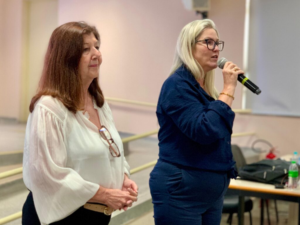 A imagem mostra duas pessoas em um ambiente interno, provavelmente durante uma conferência ou seminário. Uma delas segura um microfone e parece estar falando ao público, enquanto a outra permanece ao lado, acompanhando atentamente. Ao fundo, há uma mesa com alguns objetos, incluindo uma garrafa de água, uma bolsa preta e um extintor de incêndio, elementos que reforçam o caráter funcional do espaço.