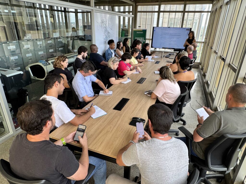 A imagem mostra uma reunião ou sessão de treinamento em um ambiente de escritório moderno e bem iluminado. Cerca de 18 pessoas estão sentadas ao redor de uma longa mesa retangular de madeira clara, enquanto uma mulher está em pé próxima a uma grande tela digital, aparentemente conduzindo a apresentação. O grupo é diverso, composto por homens e mulheres de diferentes idades e etnias, com vestimentas que vão do casual ao casual de negócios. Alguns participantes fazem anotações em cadernos, outros olham para seus celulares, e todos parecem atentos à apresentação. O espaço tem um estilo industrial, com colunas de concreto, grandes janelas que permitem a entrada de luz natural abundante, e uma divisória de vidro que mostra um espaço de armazenamento com armários metálicos. No fundo, há uma faixa verde com o texto "SECRETARIA MUNICIPAL DE HABITAÇÃO E REGULARIZAÇÃO FUNDIÁRIA", indicando que o encontro está relacionado a essa secretaria pública. A composição da imagem utiliza as linhas da mesa e das janelas para direcionar o olhar para a apresentadora, transmitindo uma atmosfera de colaboração e foco. A iluminação é uniforme e destaca os detalhes do ambiente e das pessoas presentes.