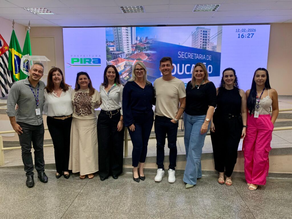 A imagem mostra um grupo de nove adultos posicionados lado a lado em um ambiente interno bem iluminado, provavelmente uma sala de conferências ou reunião oficial. Ao fundo, há uma grande tela digital exibindo uma paisagem urbana e os dizeres “SECRETARIA DE EDUCAÇÃO”, acompanhados do logotipo da Prefeitura de Piracicaba. Também aparecem na tela a data “12-02-2026” e o horário “16:27”. À esquerda, três bandeiras estão dispostas, incluindo a do Brasil e a do estado de São Paulo. Os participantes estão vestidos em trajes de estilo profissional ou casual de negócios, sugerindo que se trata de um evento institucional ou anúncio formal relacionado à área da educação.