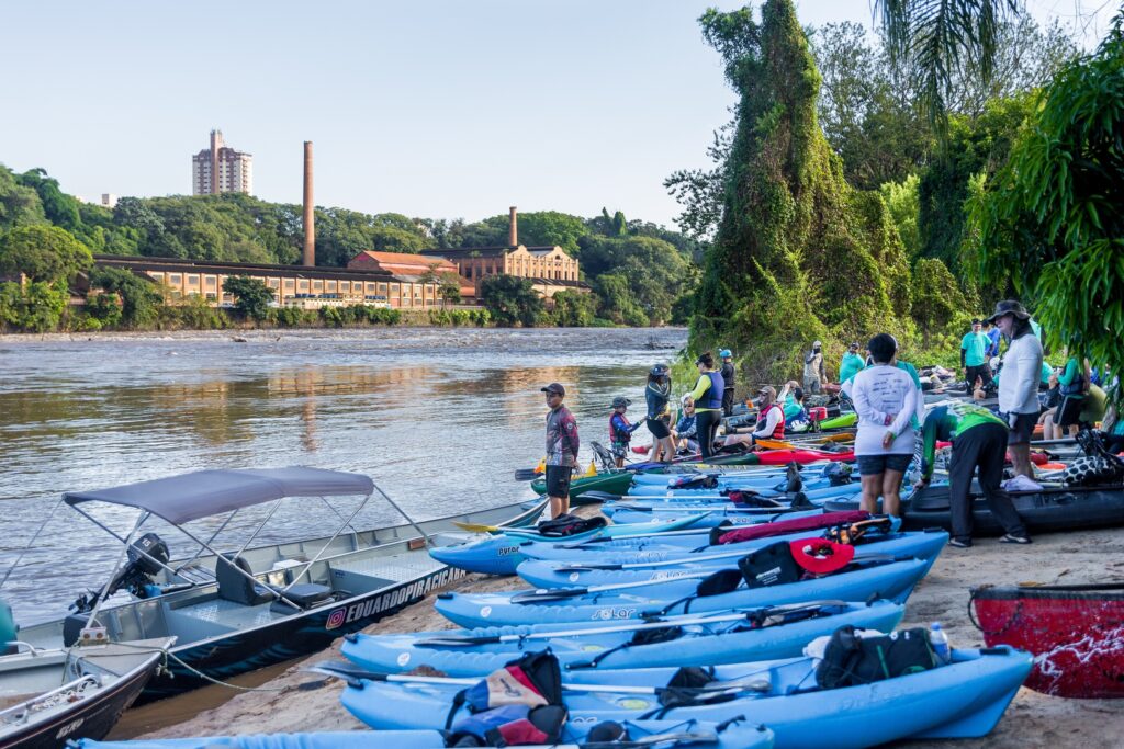 A imagem mostra um grupo de pessoas se preparando para um passeio de caiaque à beira de um rio. Na margem arenosa, há uma longa fileira de caiaques azuis claros alinhados, prontos para serem usados. À esquerda, um barco motorizado metálico está atracado, com o nome de usuário do Instagram "@EDUARDOPIRACICABA" visível na lateral. Algumas pessoas estão próximas aos caiaques, vestindo coletes salva-vidas e roupas esportivas, algumas com camisetas de marcas como "TRIMIX" e "core club". O rio tem água barrenta e corre da esquerda para a direita da imagem. Ao fundo, do outro lado do rio, há uma extensa área verde com muitas árvores, e além delas, construções industriais antigas de tijolos com chaminés altas, sugerindo uma antiga fábrica. O céu está claro, indicando um dia ensolarado. A composição da foto usa a linha dos caiaques para guiar o olhar para dentro da cena, transmitindo uma atmosfera de lazer e atividade ao ar livre.