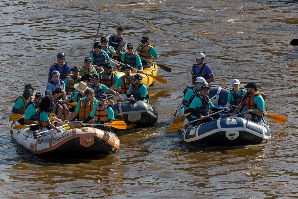 A imagem mostra um grupo de pessoas praticando rafting em um rio de águas marrons e turbulentas. São três botes infláveis coloridos: um branco e laranja à esquerda com o logo "EcoBrotas", um azul escuro no centro-direita e um amarelo ao fundo. Cada bote está cheio de pessoas vestindo coletes salva-vidas e capacetes, algumas usando camisetas azul-turquesa. O ambiente é ao ar livre, com luz do dia forte que destaca o movimento da água e as cores dos botes. A cena transmite aventura e diversão em um cenário natural, provavelmente em um local turístico de ecoturismo no Brasil.