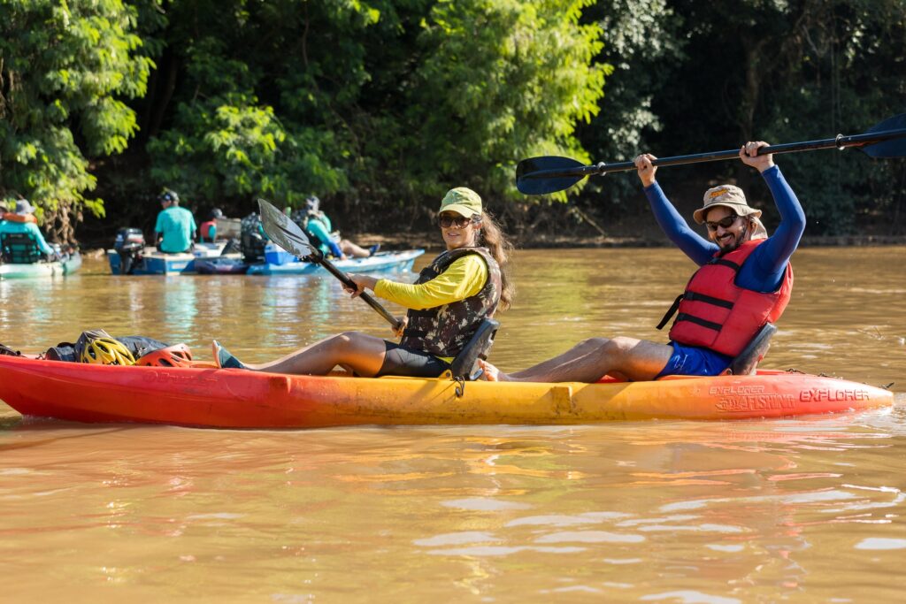 A imagem mostra duas pessoas em um caiaque duplo, navegando por um rio de água turva e marrom. O caiaque é amarelo com detalhes em laranja e tem a inscrição "EXPLORER FISHING" na lateral. A mulher, que está na frente, veste uma camisa amarela de manga longa, um colete salva-vidas camuflado, um boné verde e óculos escuros, enquanto segura um remo preto. O homem, que está atrás, usa uma camisa azul de manga longa, colete salva-vidas vermelho, shorts azuis, chapéu bege e óculos escuros; ele sorri e levanta o remo acima da cabeça. Ao fundo, há uma densa floresta verde e outros caiaques também no rio, sugerindo um passeio recreativo ou uma expedição ao ar livre em um dia ensolarado. A atmosfera é de aventura e contato com a natureza.