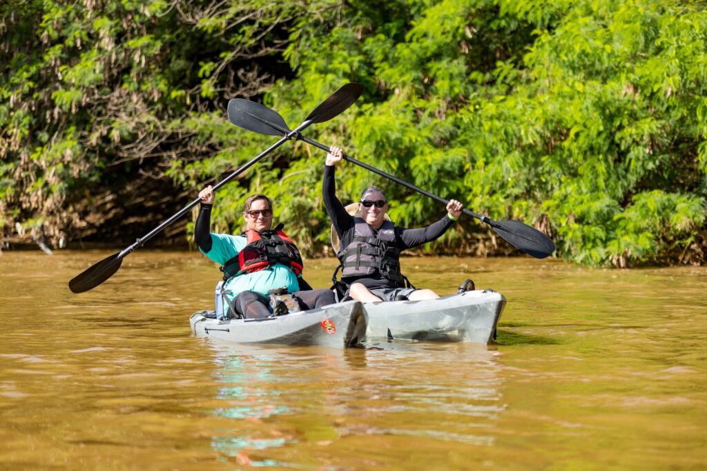 A imagem mostra dois homens praticando caiaque em um rio de água marrom. Ambos estão sentados em caiaques cinzas do tipo sit-on-top, usando coletes salva-vidas e óculos escuros. Eles estão sorrindo e levantando os remos no ar em um gesto de celebração ou vitória. O cenário ao fundo é composto por árvores e vegetação densa, sugerindo que estão em uma área natural, talvez uma floresta ou parque. A luz do dia ilumina a cena, destacando as cores vibrantes da natureza ao redor. A água tem uma tonalidade turva, característica comum em rios com bastante sedimento. A imagem transmite uma sensação de aventura, diversão e contato com a natureza.
